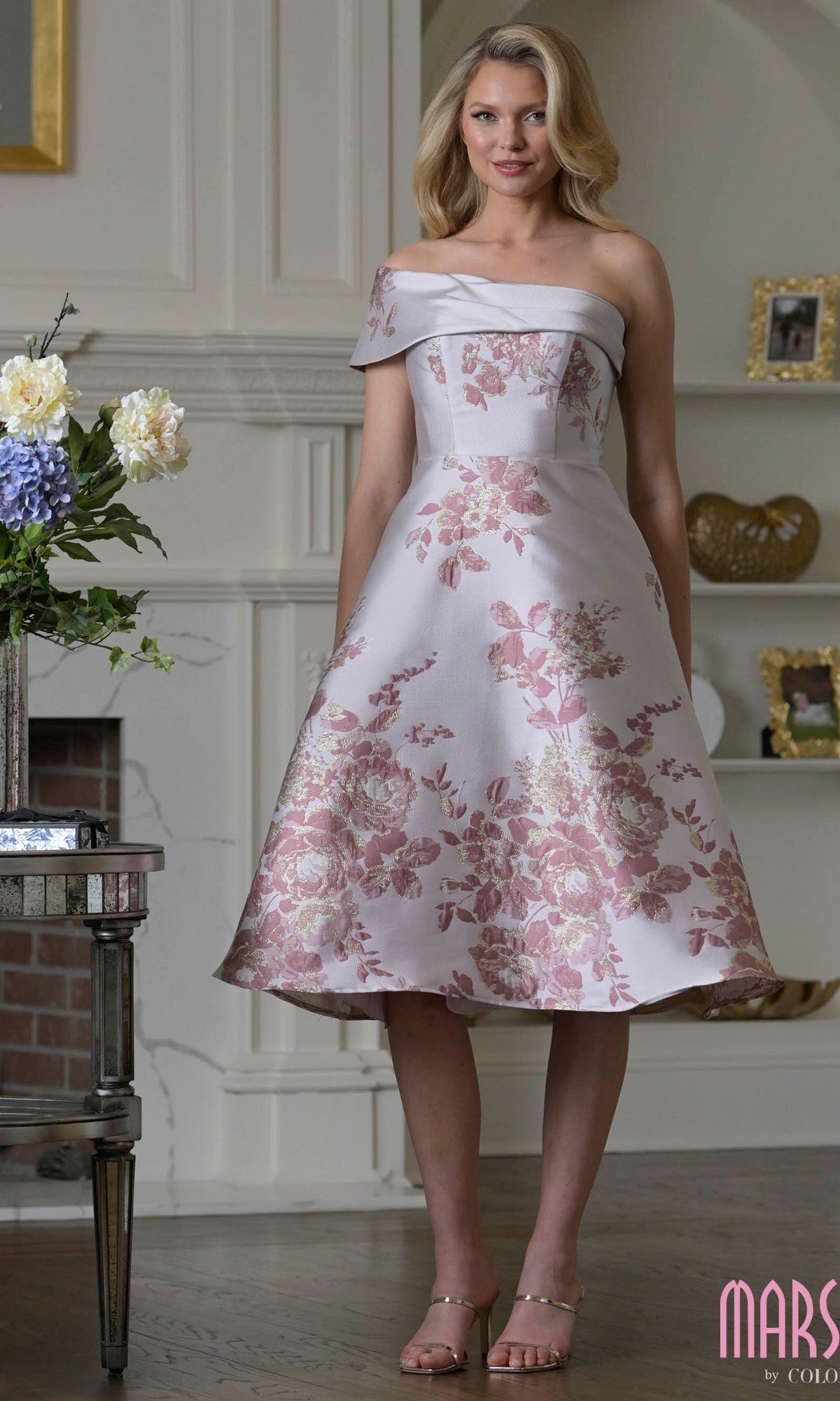 A woman stands indoors by a fireplace and a vase of flowers, wearing the Marsoni MV1415 Knee Length Semi-Formal Dress in Rose Multi.